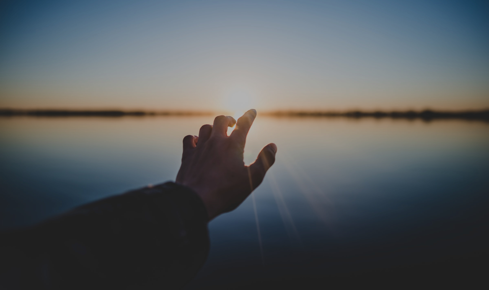A hand stretching out to reach the sun setting on a lake